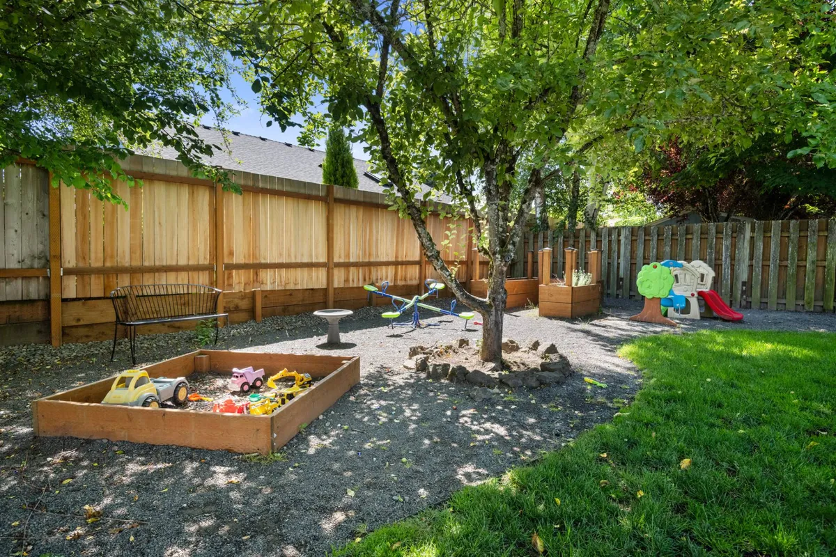 A joyful backyard scene showing a play structure with swings, a sandbox, and lush grass under the shade of a tree. The setup is clearly geared toward children, providing dedicated play areas that invite laughter and safe recreation.