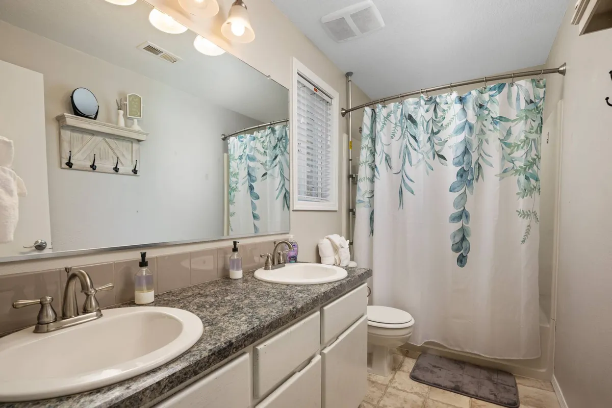 A spacious bathroom featuring double vanity sinks, a large mirror, and a shower curtain with a foliage design. The setup is practical for multiple guests and exudes a clean, inviting style.