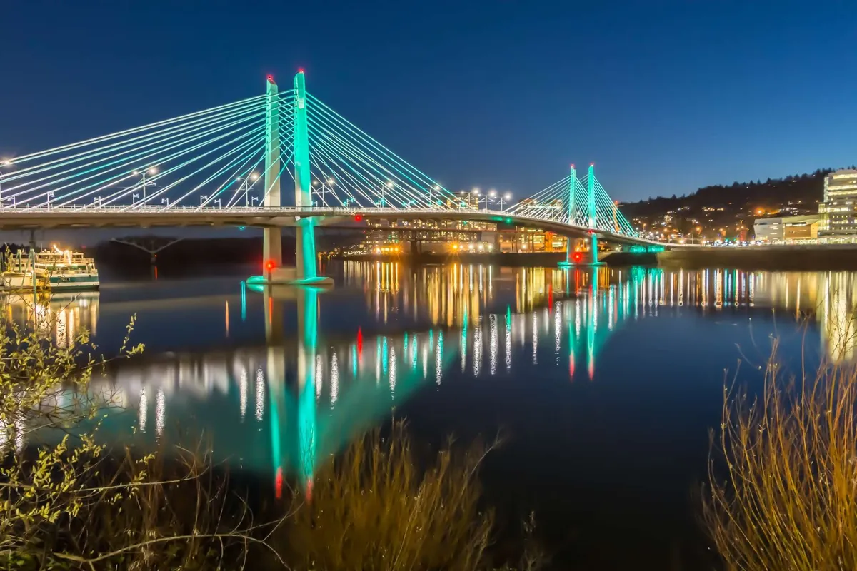 A tranquil exterior scene featuring an illuminated bridge casting reflections on calm water, with city lights twinkling in the background. The overall mood is serene and inspiring, evoking the feeling of exploring vibrant local surroundings in the evening.
