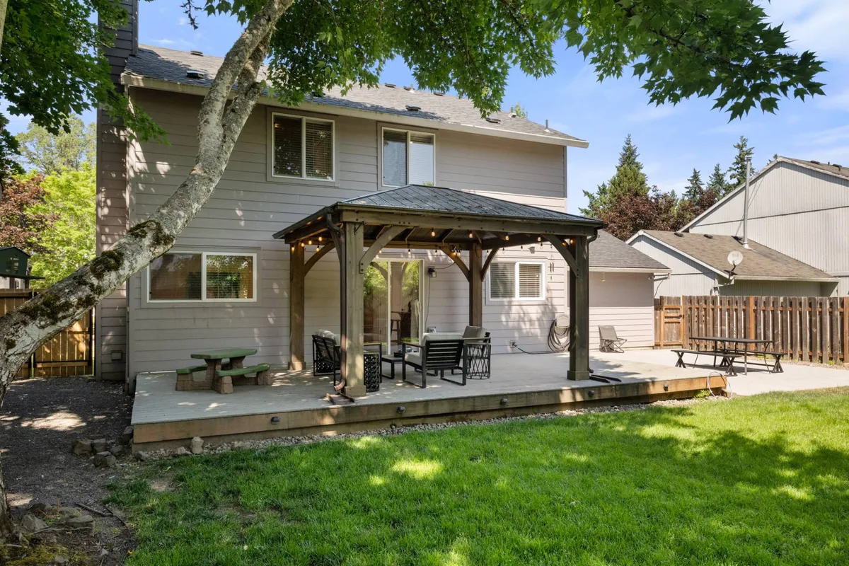 This image showcases a covered patio with comfortable outdoor furniture overlooking a green lawn. The patio is designed for relaxation, outdoor dining, and gathering, with string lights adding a cozy touch. Trees and open space make the backyard atmosphere inviting and serene.