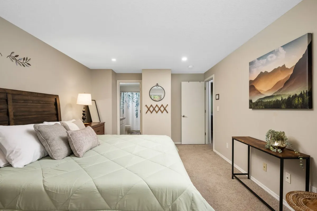 A tranquil bedroom featuring a bed with a green quilt, a nightstand with a lamp, and framed landscape artwork on the wall. Natural light streams through a window, creating a calm and inviting atmosphere.
