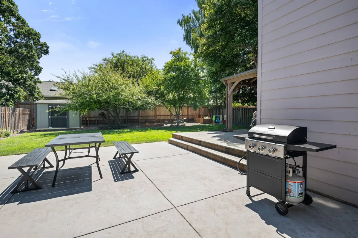 This image captures a spacious backyard scene featuring a BBQ grill beside a large outdoor dining table set atop a patio. Steps lead down to a green, well-maintained lawn that offers plenty of open space for children and pets to play. The area is framed by secure fencing, providing safety and privacy.