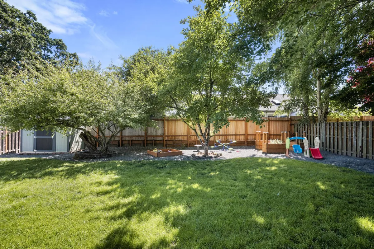 This image highlights a vibrant playground area bordered by a fence, with colorful equipment, a sandbox, lush grass, and mature trees creating ample shade. The background shows a residential setting, reinforcing the neighborhood's family-friendly atmosphere.