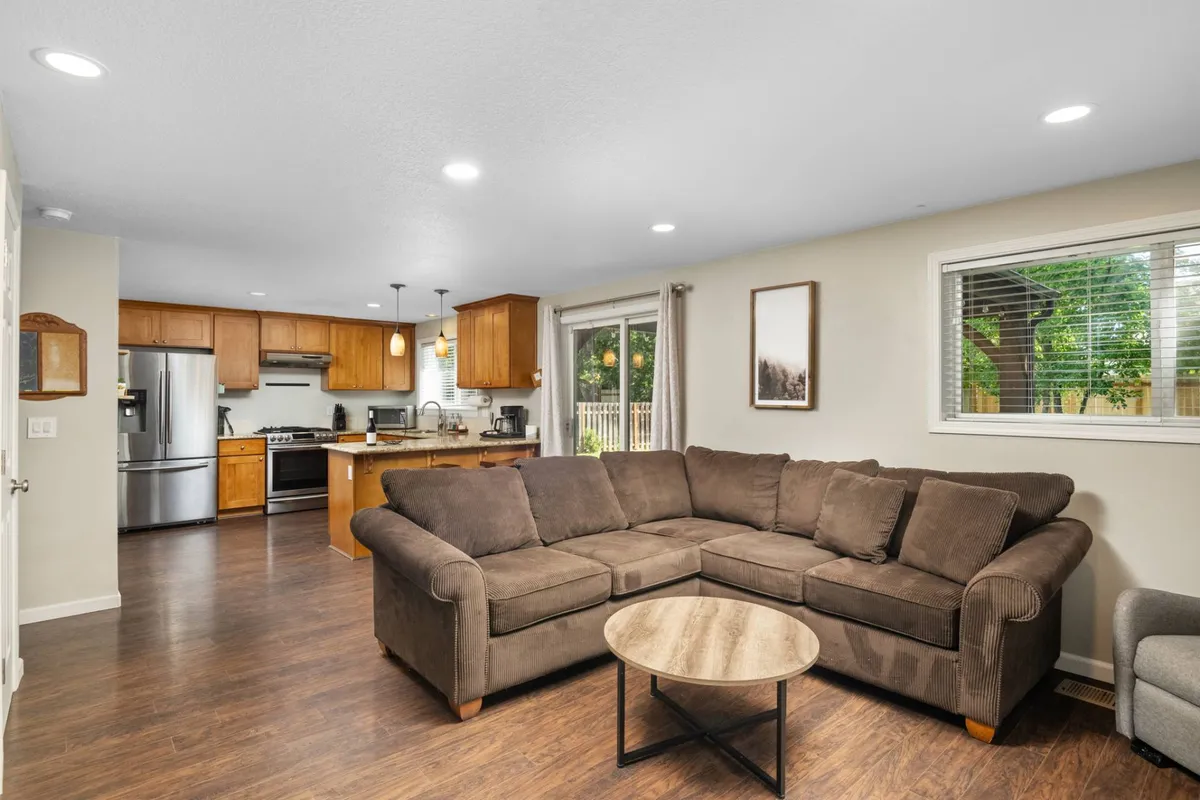 This image showcases a spacious and welcoming living area with a comfortable sectional sofa, a coffee table, and a kitchen area visible in the background. Large windows allow natural light to flood the room, creating an inviting atmosphere ideal for family gatherings and intimate conversations.