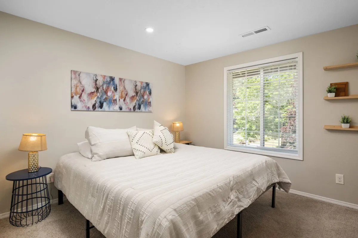 A serene bedroom space featuring a bed with decorative pillows, soft natural light streaming through windows, and lush plants on shelving, creating a calming and restorative atmosphere.