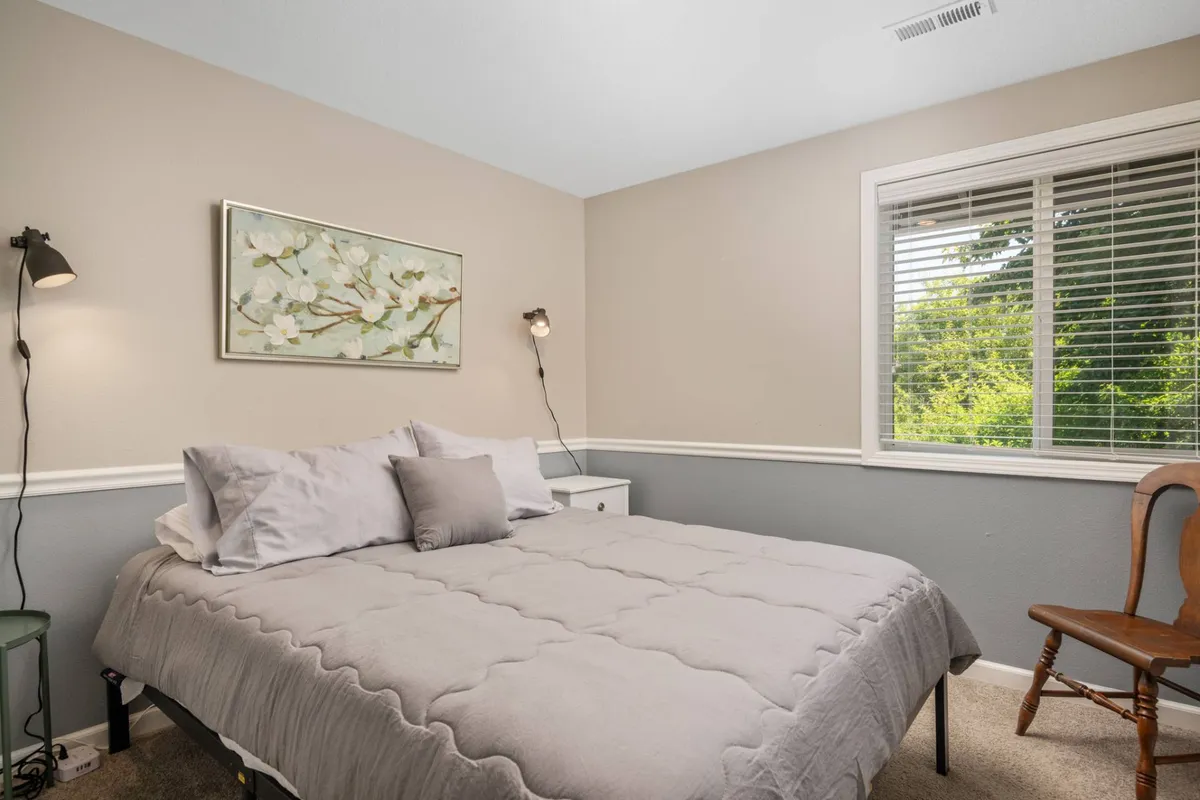 A serene bedroom with gray bedding, floral wall art, and a window letting in natural sunlight. The space feels airy and calm, with a focus on comfort and modern simplicity.
