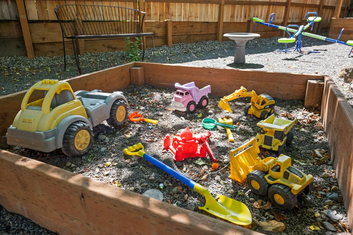 A joyful outdoor scene featuring a sandbox filled with toy vehicles and play tools, and a seesaw, all set on green grass. The area is clearly designed for imaginative play and social interactions among children.
